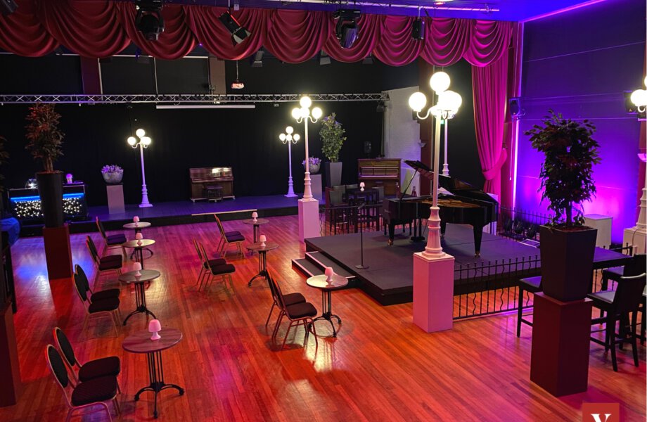 Ruime zaal met podium, lantarenpalen en groepjes van twee stoelen met een kleine tafel